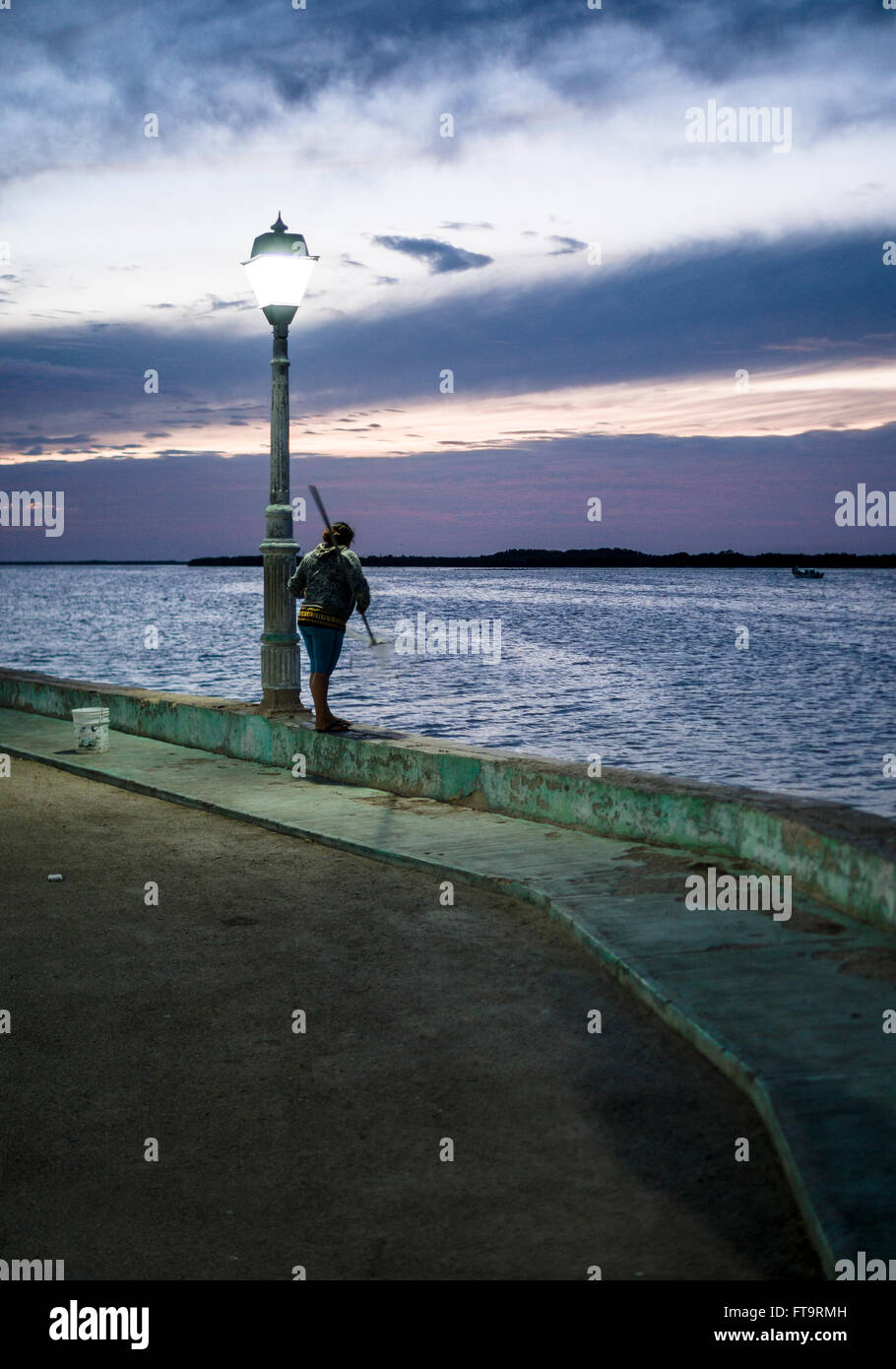 L'écopage des crevettes au crépuscule sur le mur de la mer à Rio Lagartos. Une rue le témoin reste allumé avec un ciel de coucher du soleil comme un pêcheur de crevettes tourne son filet Banque D'Images