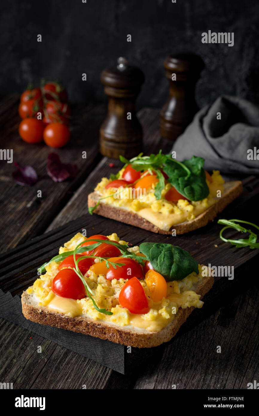 Toast pour le petit déjeuner avec des œufs brouillés, fromage, tomates cerises, roquette et salade de maïs sur fond de bois rustique Banque D'Images