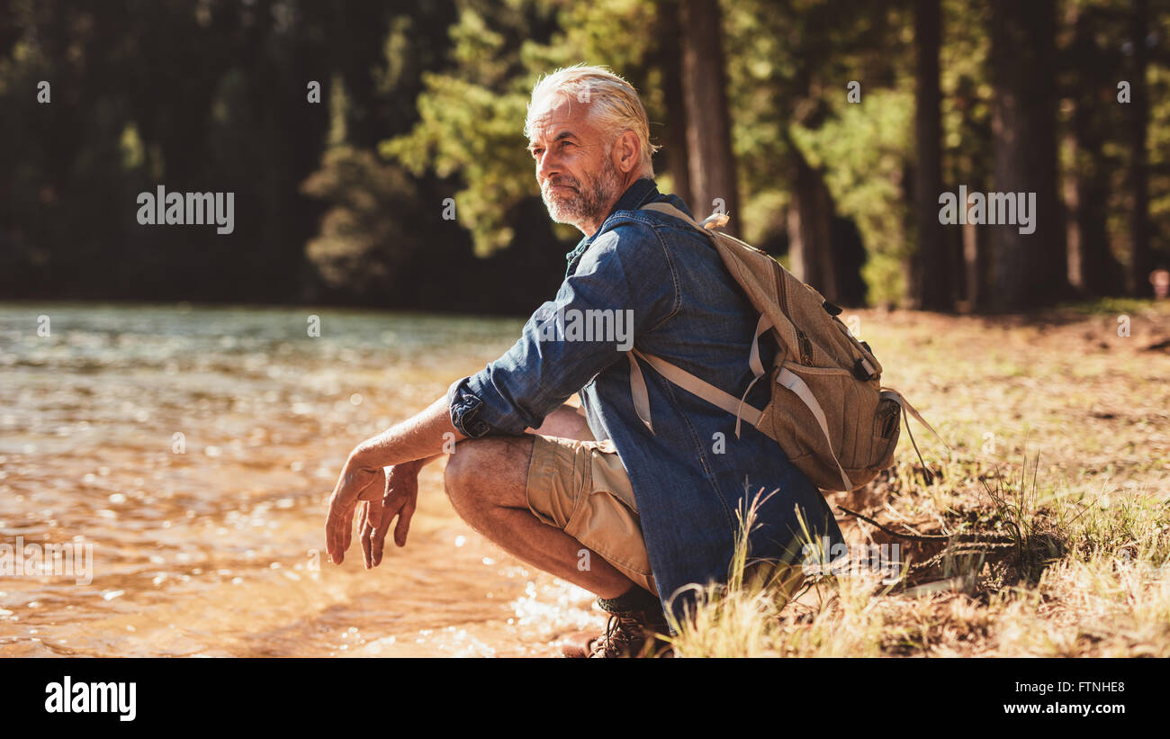 Portrait of a senior man with backpack assis près d'un lac en admirant la vue. Homme mûr en faisant une pause de sa randonnée pédestre et loo Banque D'Images