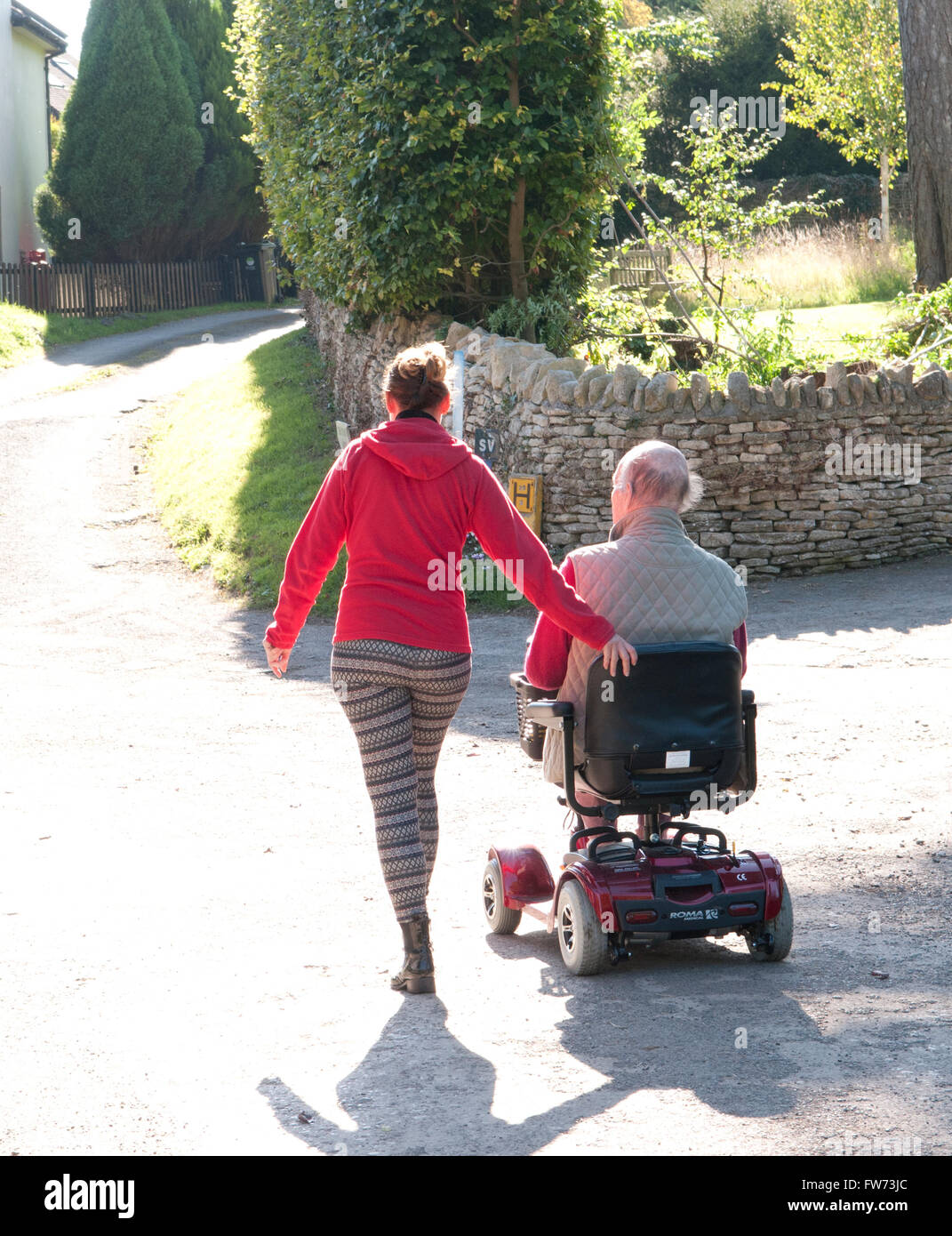 Vue arrière d'un homme âgé à l'aide d'un scooter de mobilité avec son soignant marcher à côté de lui Banque D'Images