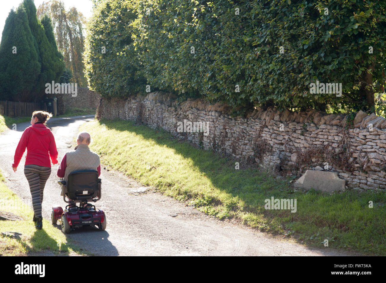 Vue arrière d'un homme âgé à l'aide d'un scooter de mobilité avec son soignant marcher à côté de lui Banque D'Images