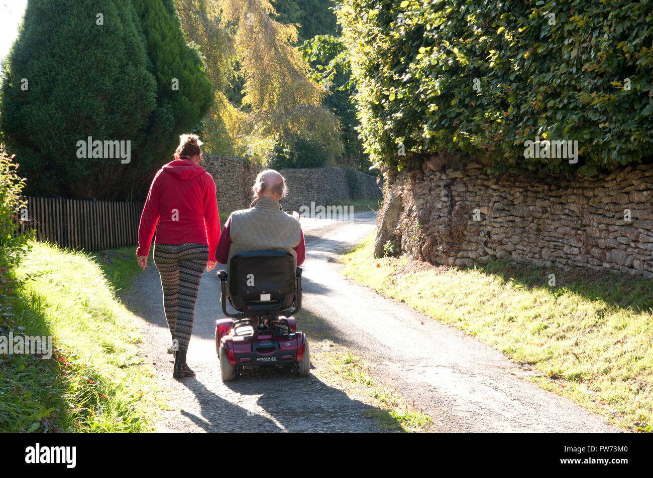 Vue arrière d'un homme âgé à l'aide d'un scooter de mobilité avec son soignant marcher à côté de lui Banque D'Images