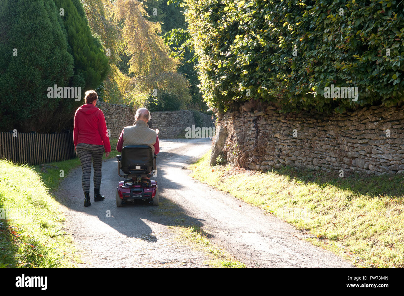 Vue arrière d'un homme âgé à l'aide d'un scooter de mobilité avec son soignant marcher à côté de lui Banque D'Images
