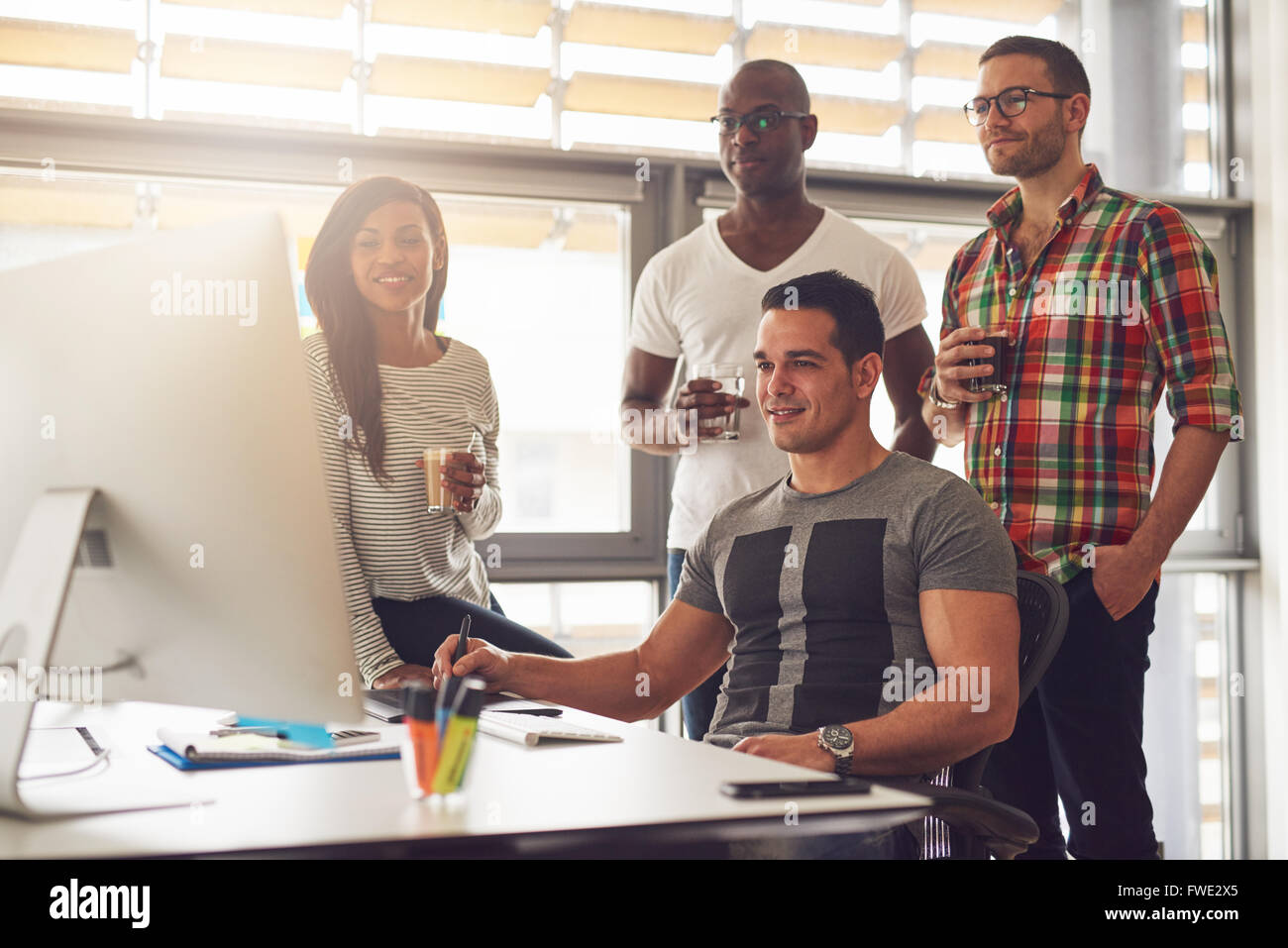 Quatre jeunes détendue mâle et femelle divers travailleurs de petites entreprises avec des boissons dans la main debout autour de computer in office Banque D'Images