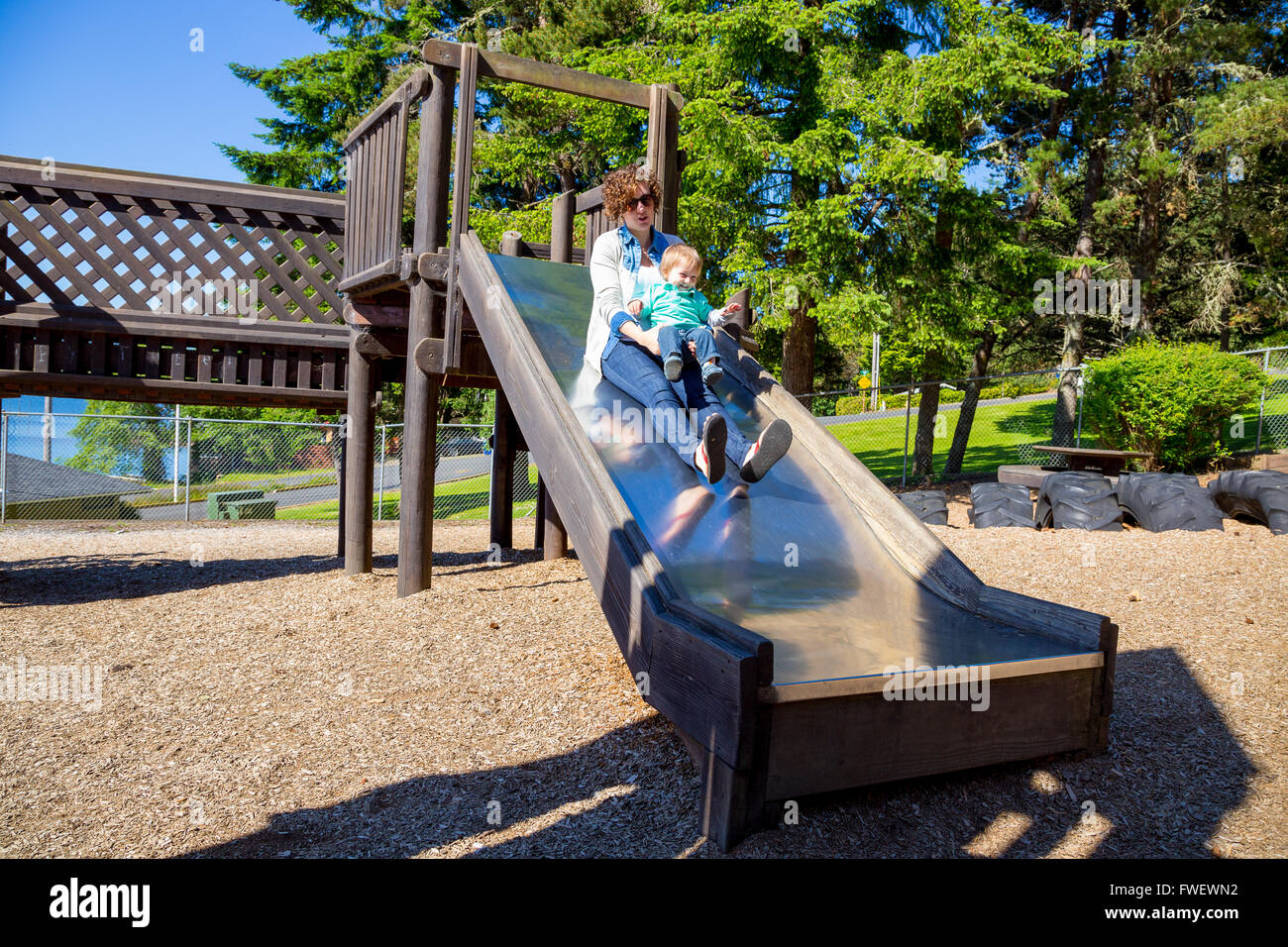 Une maman et son fils glisser et tomber dans une diapositive de métal à une aire de jeux extérieure dans un parc. Banque D'Images