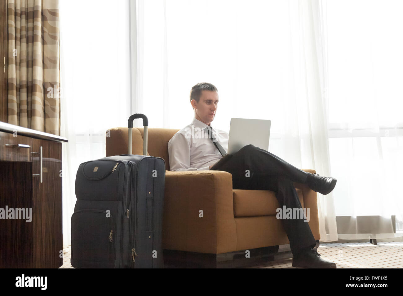 Portrait of handsome young man wearing white shirt officiel cravate noire et assis dans un fauteuil dans chambre d'hôtel avec paniers Banque D'Images