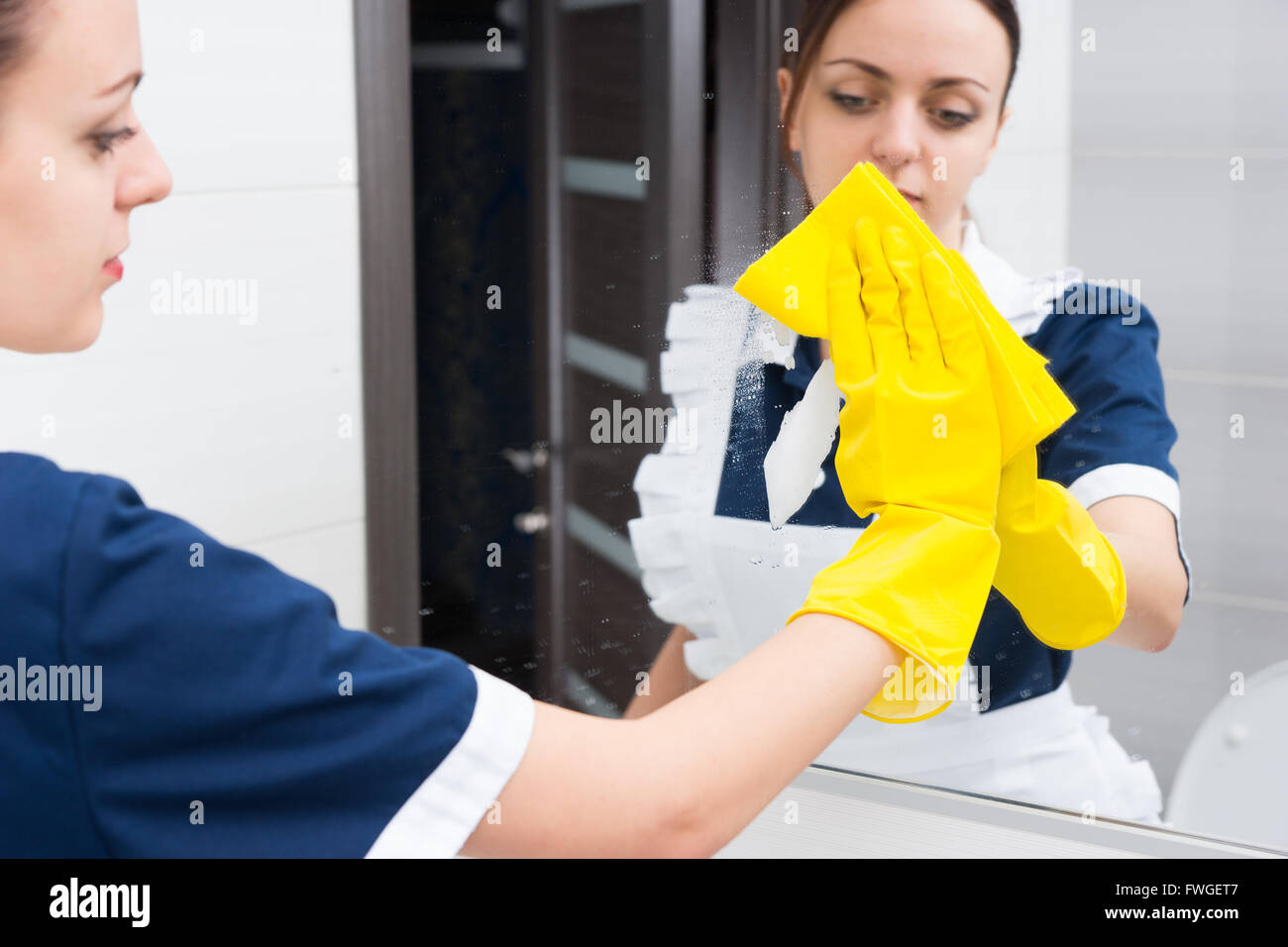 Reflet de la jeune femme adulte sérieux hotel maid à l'aide d'une éponge jaune et des gants en caoutchouc pour nettoyer le miroir tout en travaillant dans une salle de bains privative. Banque D'Images