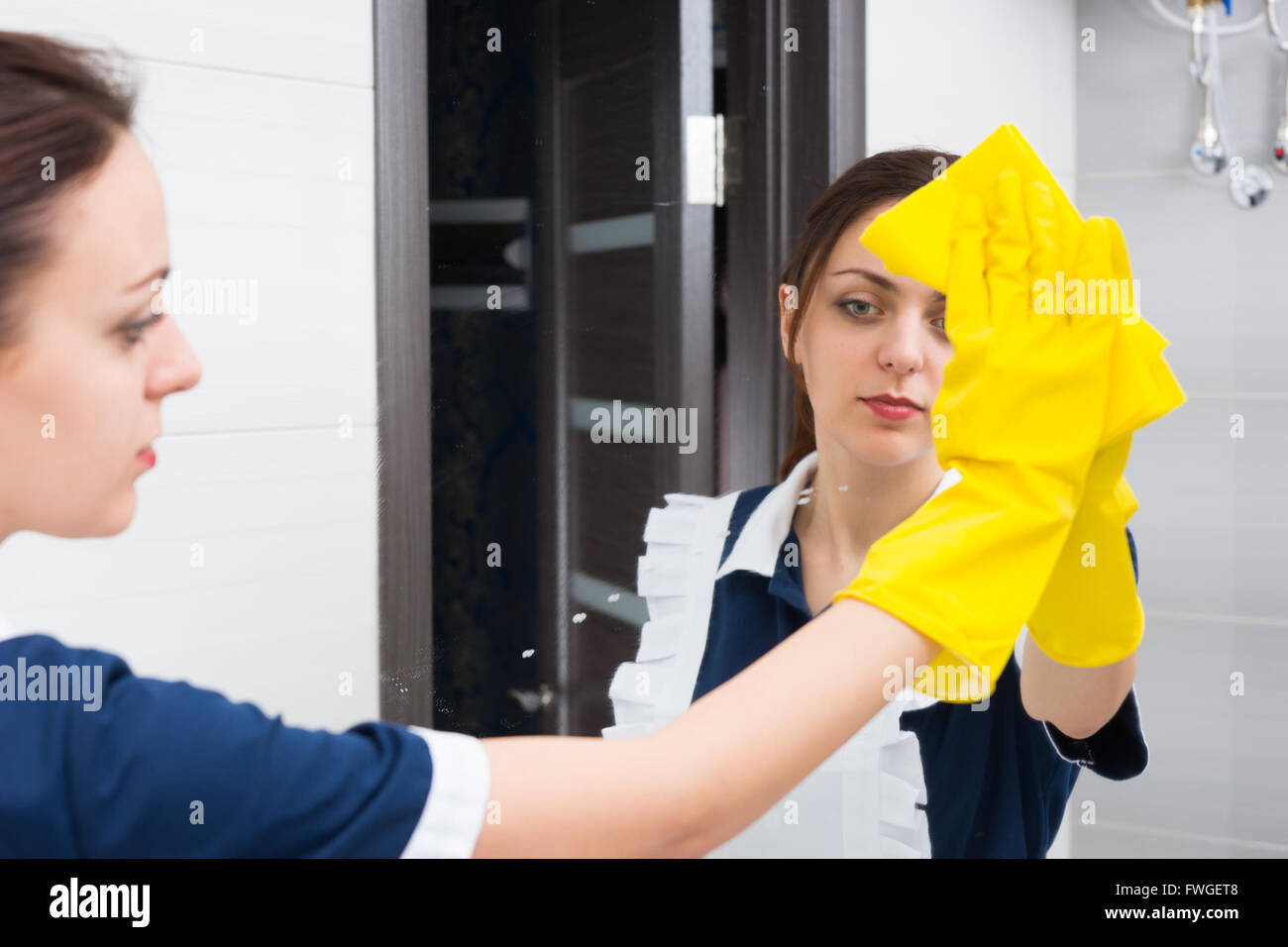 La réflexion de calme jeune femelle adulte portrait à l'aide d'une éponge jaune et des gants en caoutchouc pour nettoyer un miroir à main tout en travaillant dans une salle de bains privative. Banque D'Images