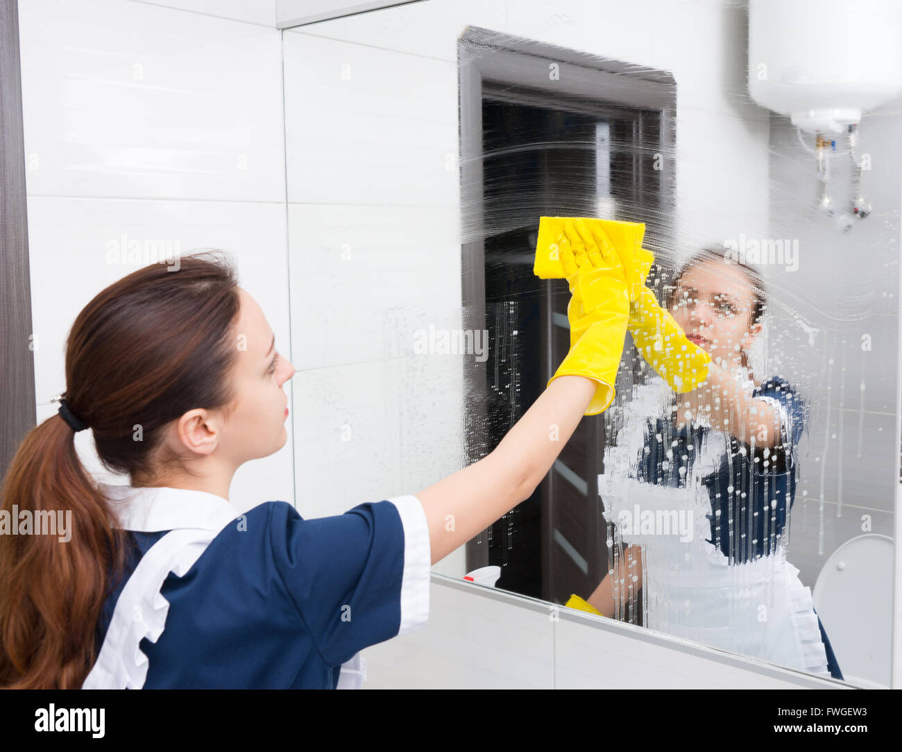 Miroir de salle de bains hôtel humide étant essuyer avec un chiffon jaune par maid en uniforme bleu et blanc et jaune des gants en caoutchouc Banque D'Images