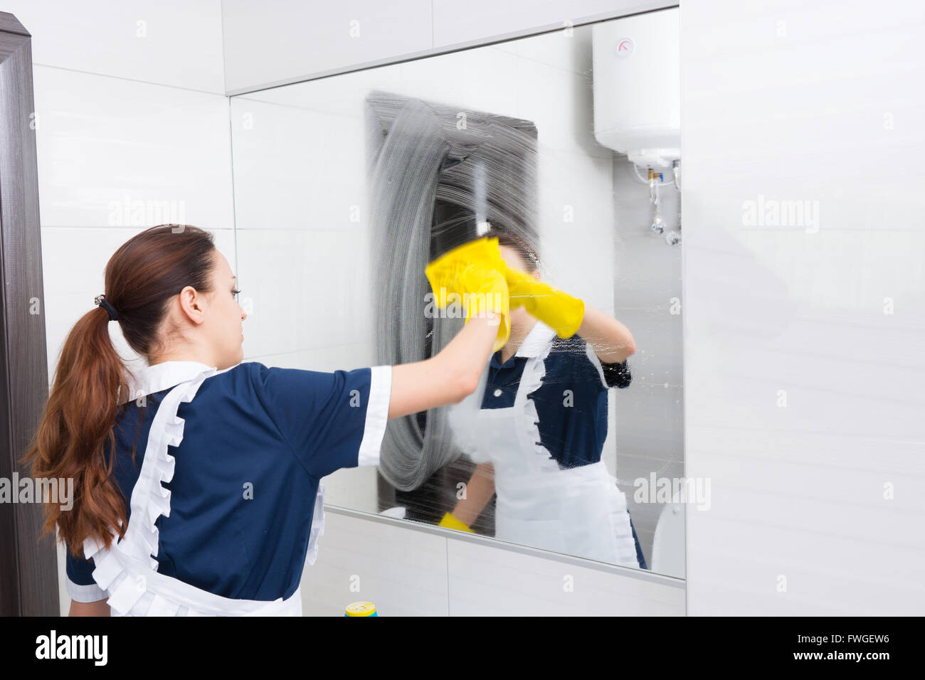 Hôtel Grand miroir de salle de bains enduite avec du détergent en cours de nettoyage avec chiffon jaune par maid en uniforme bleu et blanc et jaune des gants en caoutchouc. Banque D'Images