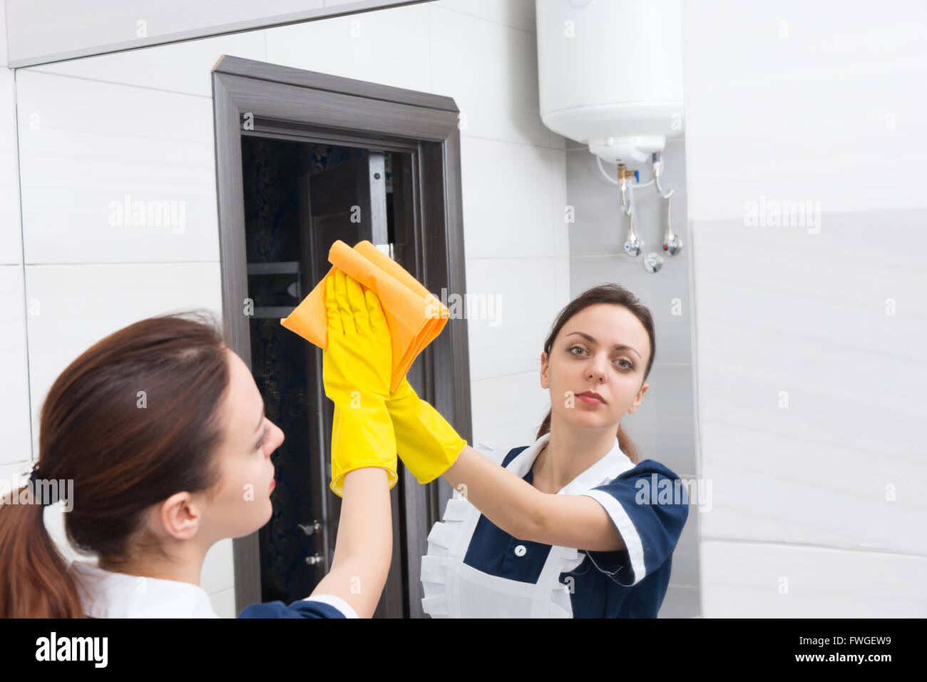 Jeune femme sérieuse femme portant des gants de caoutchouc à la recherche de miroir tandis qu'une salle de bains privative avec chiffon de nettoyage jaune Banque D'Images