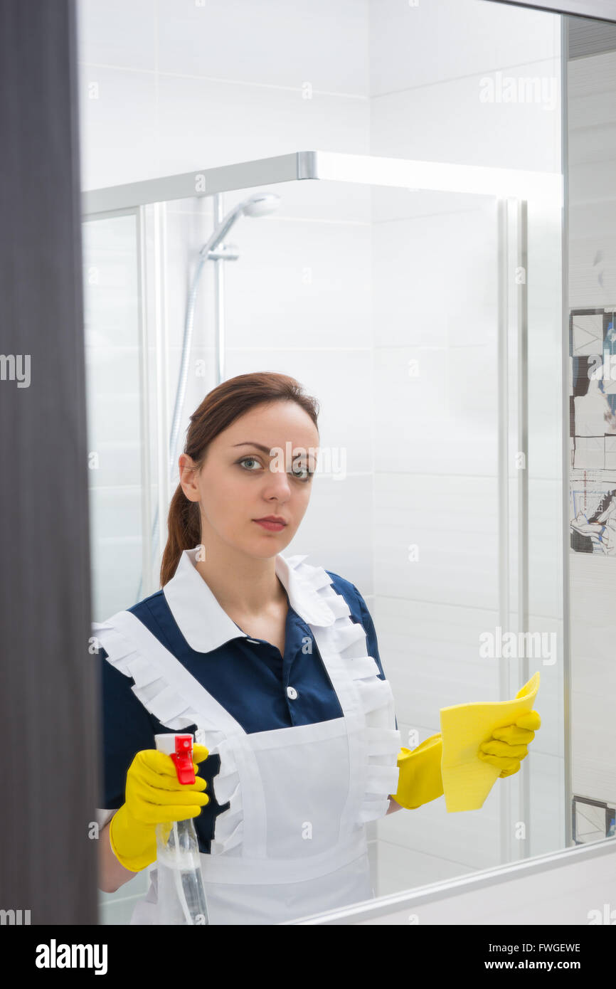 Seul cute young maid avec sérieux à la recherche dans le miroir tout en maintenant un tissu clair vaporisateur et en jaune les mains gantées. Banque D'Images