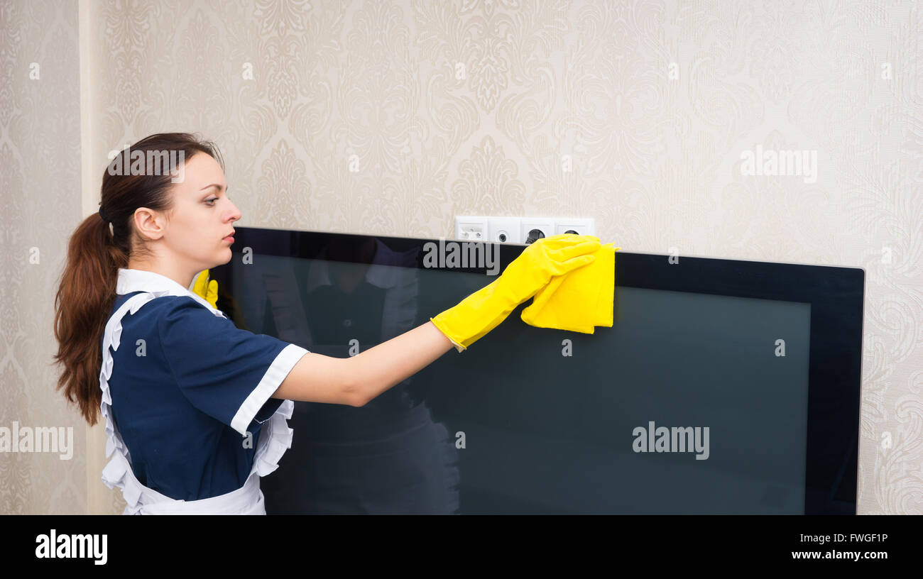 Femme de ménage ou une aide ménagère nettoyage d'un appareil de télévision de poussière le dessus avec un chiffon jaune vêtu d'un uniforme propre, profil haut du corps. Banque D'Images