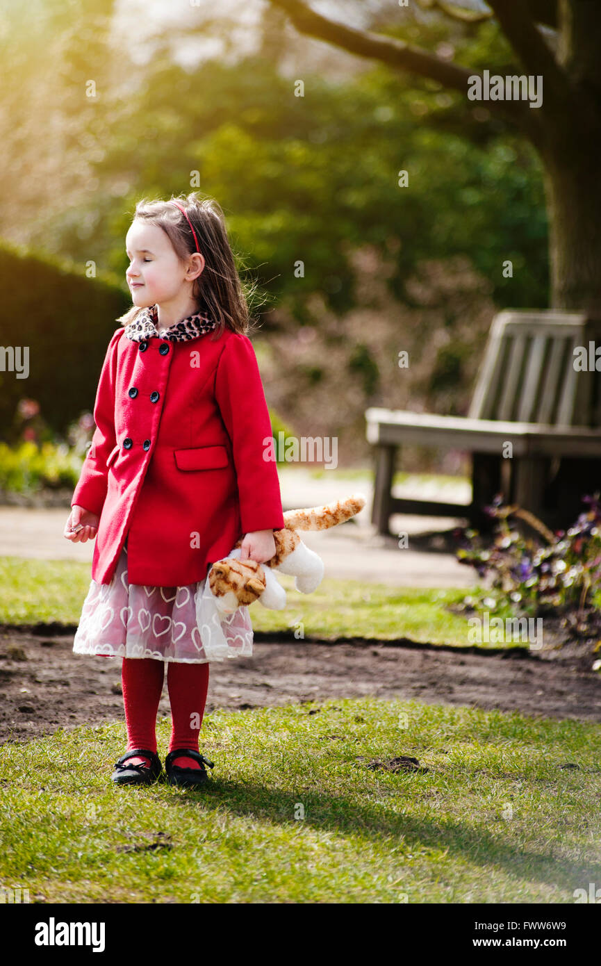 Une petite fille perdue wearing red coat standing in park holding a toy Banque D'Images