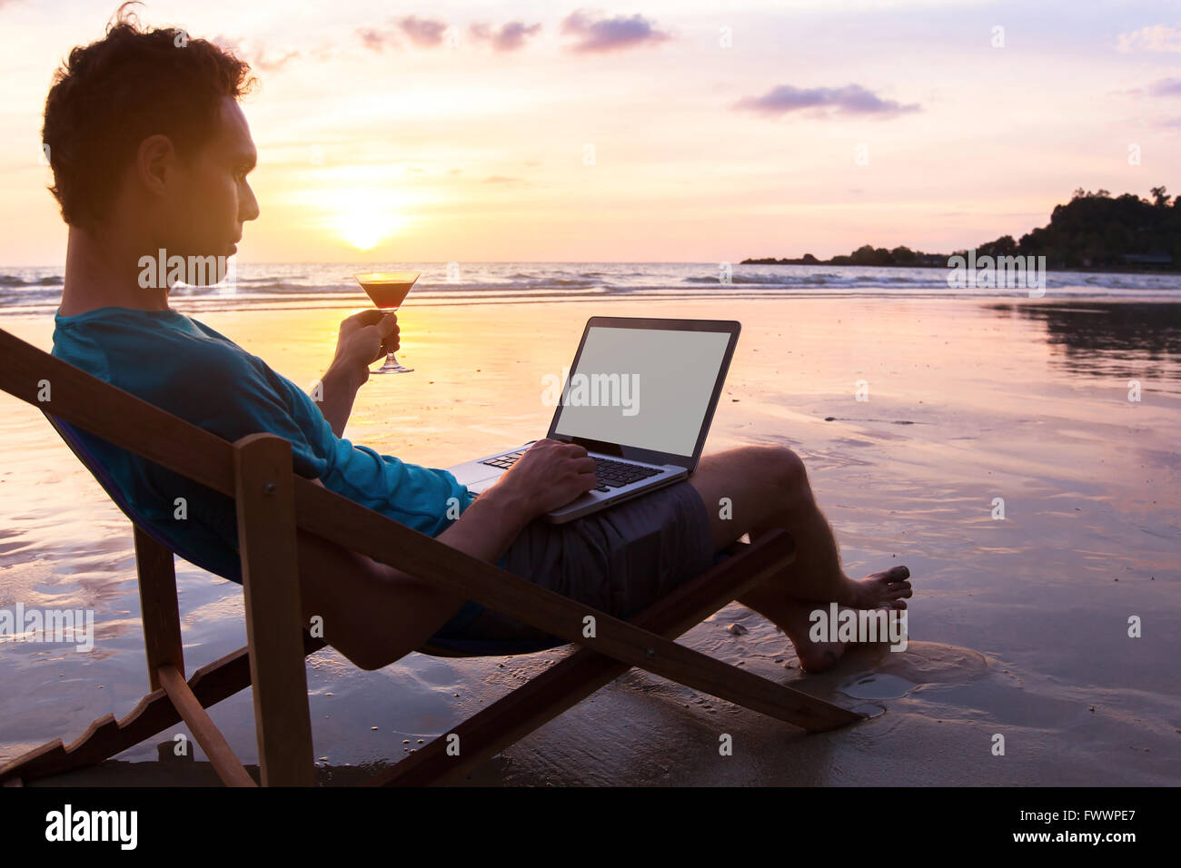 Jeune homme d'affaires avec cocktail de travailler sur un ordinateur portable sur la plage au coucher du soleil, des offres d'emploi en ligne, l'accent sur l'écran Banque D'Images