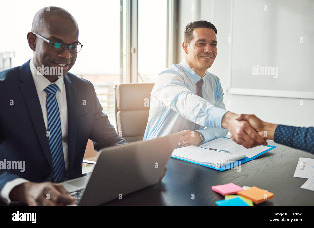 Smiling young Hispanic business man and woman shaking hands à travers une table dans le bureau vu par un smiling African American m Banque D'Images