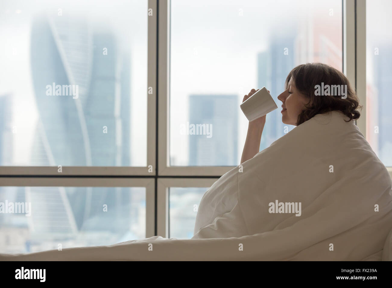 Vue de profil de jeune femme assise sur le lit défait après le réveil et tasse de café potable. Modèle féminin à la maison Banque D'Images
