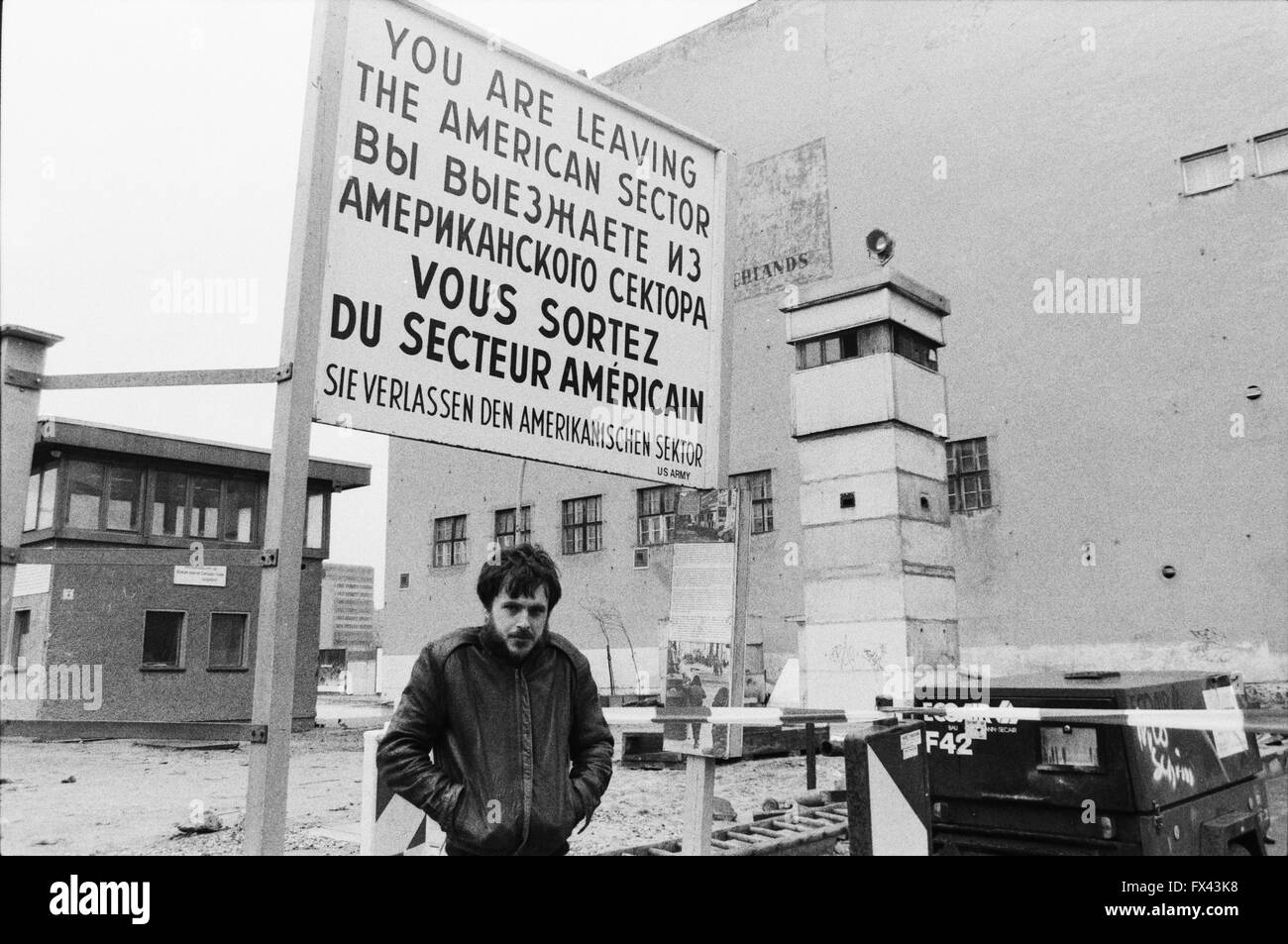 Image d'archive d'un homme en manteau de cuir debout près du Checkpoint Charlie panneau près de la Potsdamer Platz, Berlin, Allemagne, Mars 1994 "Vous quittez le secteur américain" "Vous sortez du secteur américain' 'Sie verlassen den Amerikanischen sektor', avec tour de guet. Parution du modèle. Banque D'Images