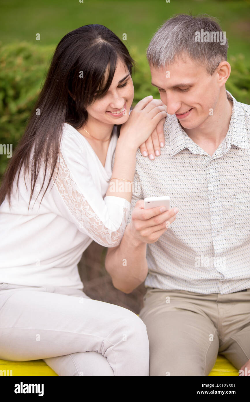 Young attractive happy smiling couple amoureux amoureux assis sur le banc de parc à ce jour, s'étreindre, holding cellphone Banque D'Images