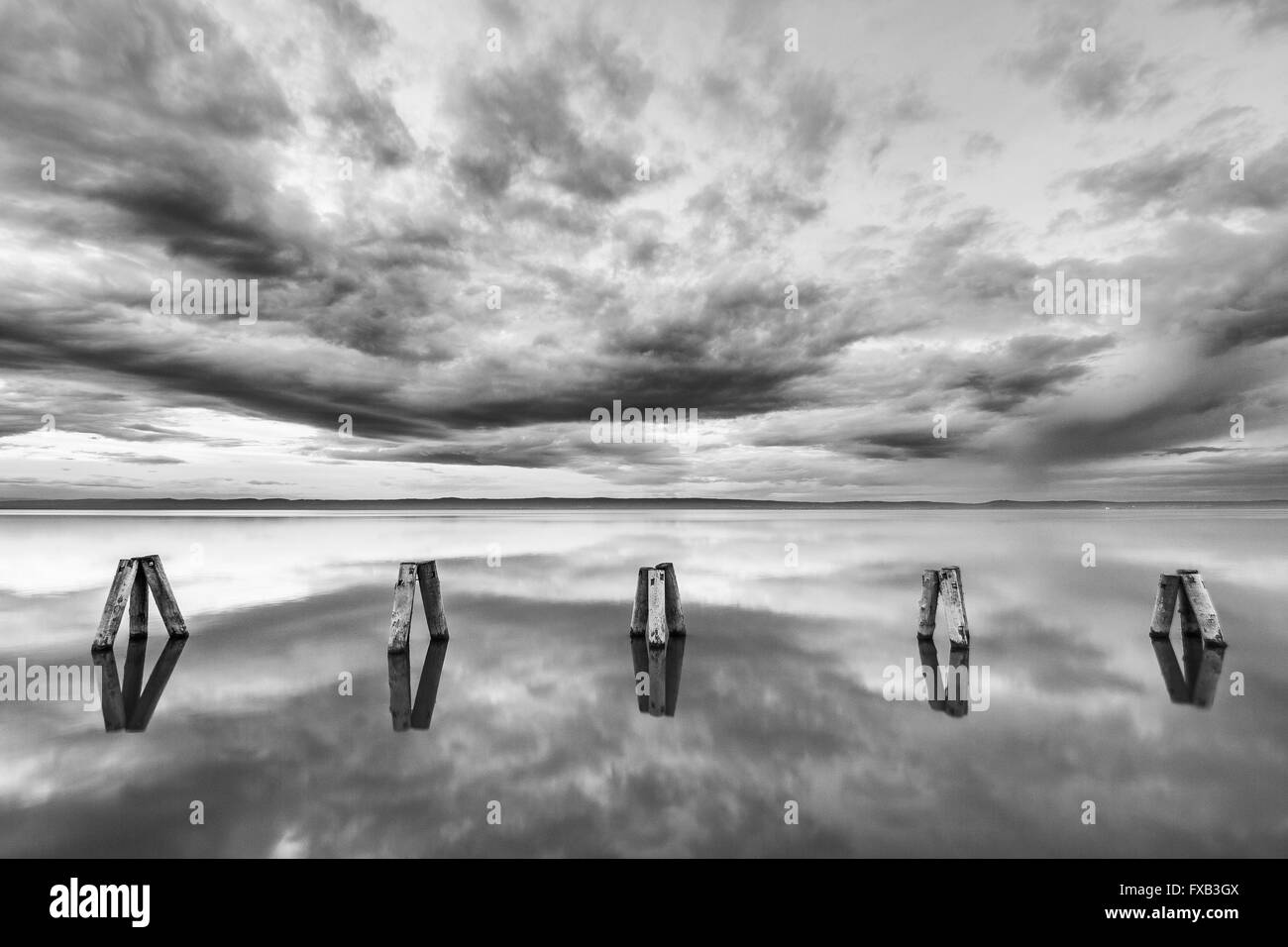 Piliers de bois reflètent dans le lac encore sur un ciel nuageux en soirée. Photo en noir et blanc. Banque D'Images