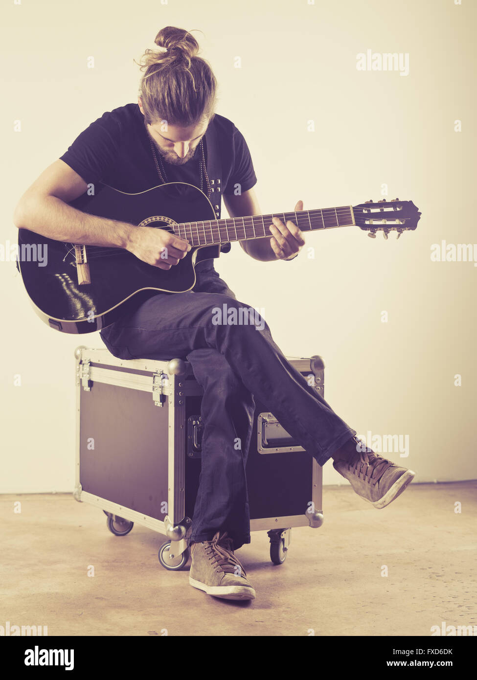 Photo d'un jeune homme séduisant aux cheveux longs et la barbe assis sur un cas de vol et en jouant une guitare acoustique. Filtré à lo Banque D'Images