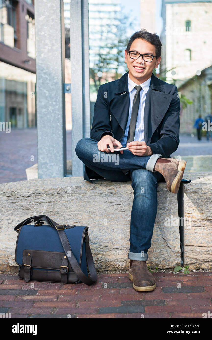 Young smiling asian business man looking at camera holding mobile phone assis dehors on city street Banque D'Images