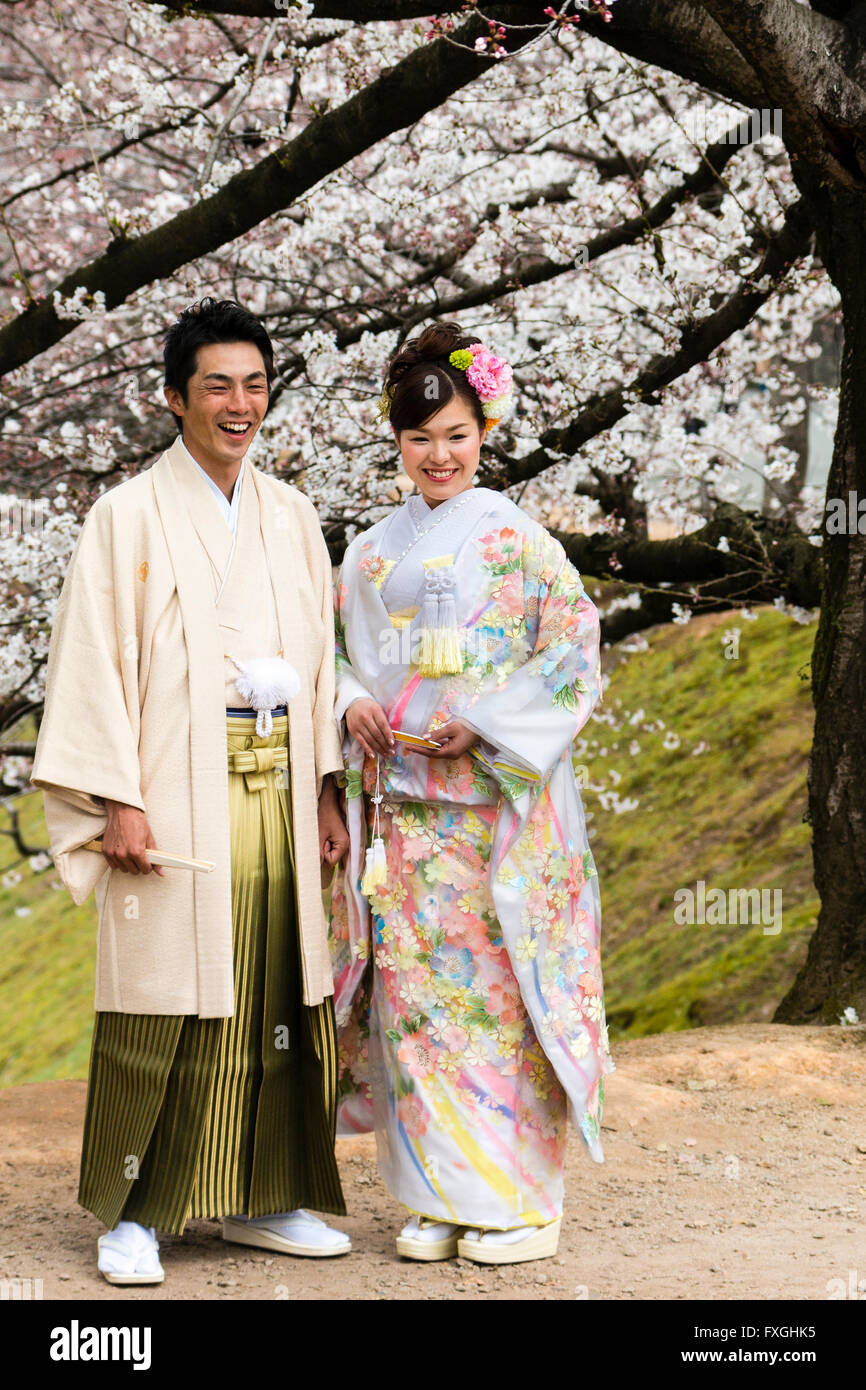 Jeune couple japonais. L'homme et de la femme, mariés en tenue de cérémonie, en face, mais pas de contact visuel, les deux rient, heureux. Les fleurs de cerisier derrière eux. Banque D'Images