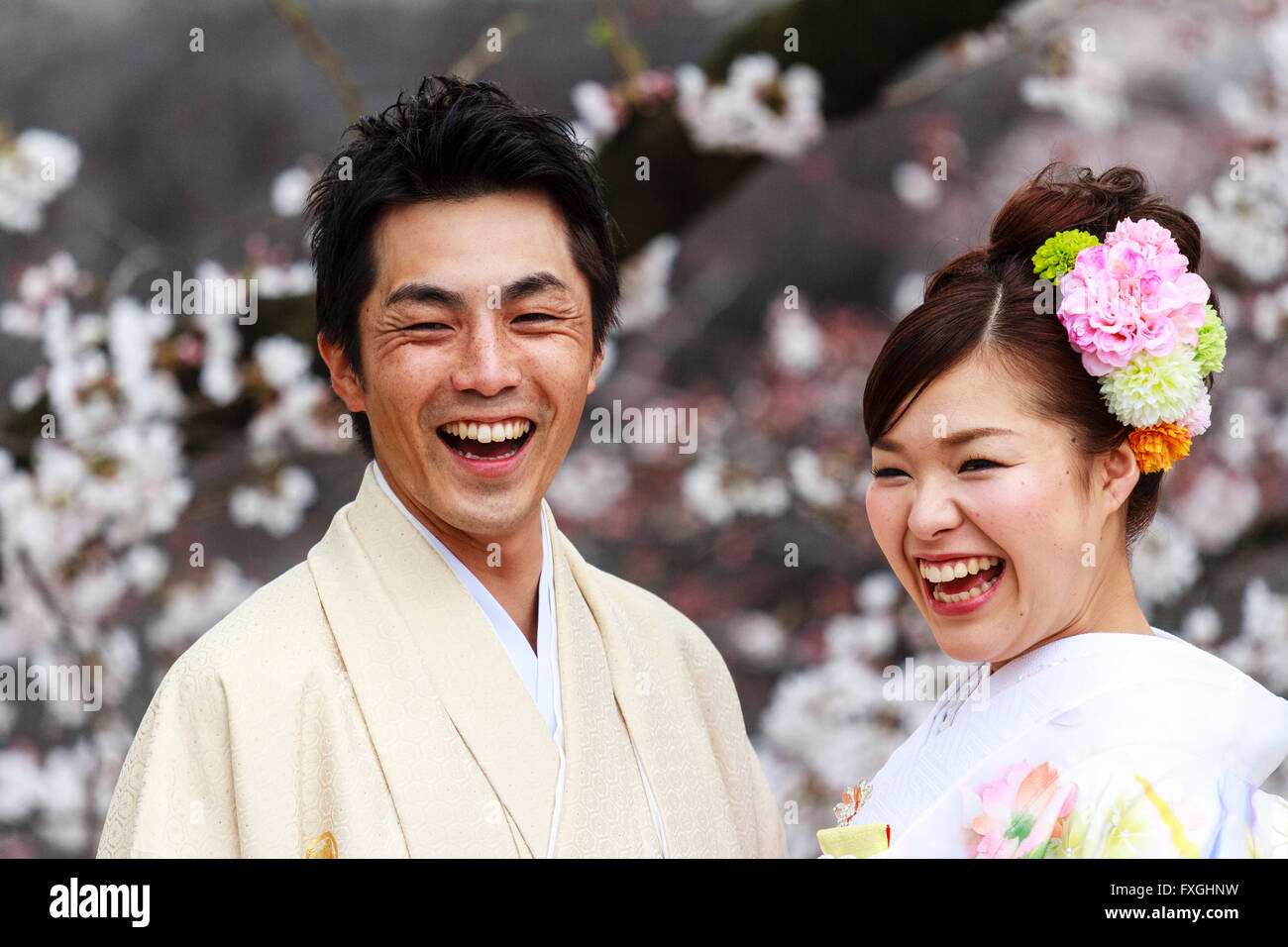 Jeune couple japonais. L'homme et de la femme, mariée et le marié. Tête et épaules, usure formelle, en face, les deux rient, heureux. Les fleurs de cerisier derrière eux. Banque D'Images
