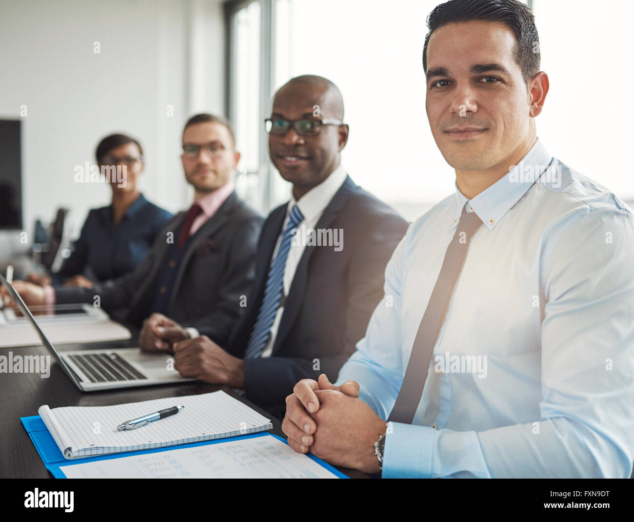 Calme groupe diversifié de quatre hommes d'affaires assis à table avec ordinateur et portables en face de grande fenêtre de bureau avec lig Banque D'Images