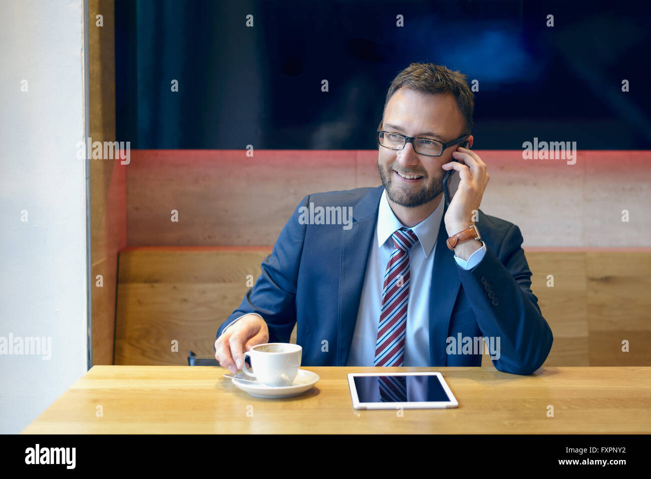 Handsome businessman avec cravate rayée est assis à table avec café et d'appels téléphoniques en utilisant sa tablette Banque D'Images