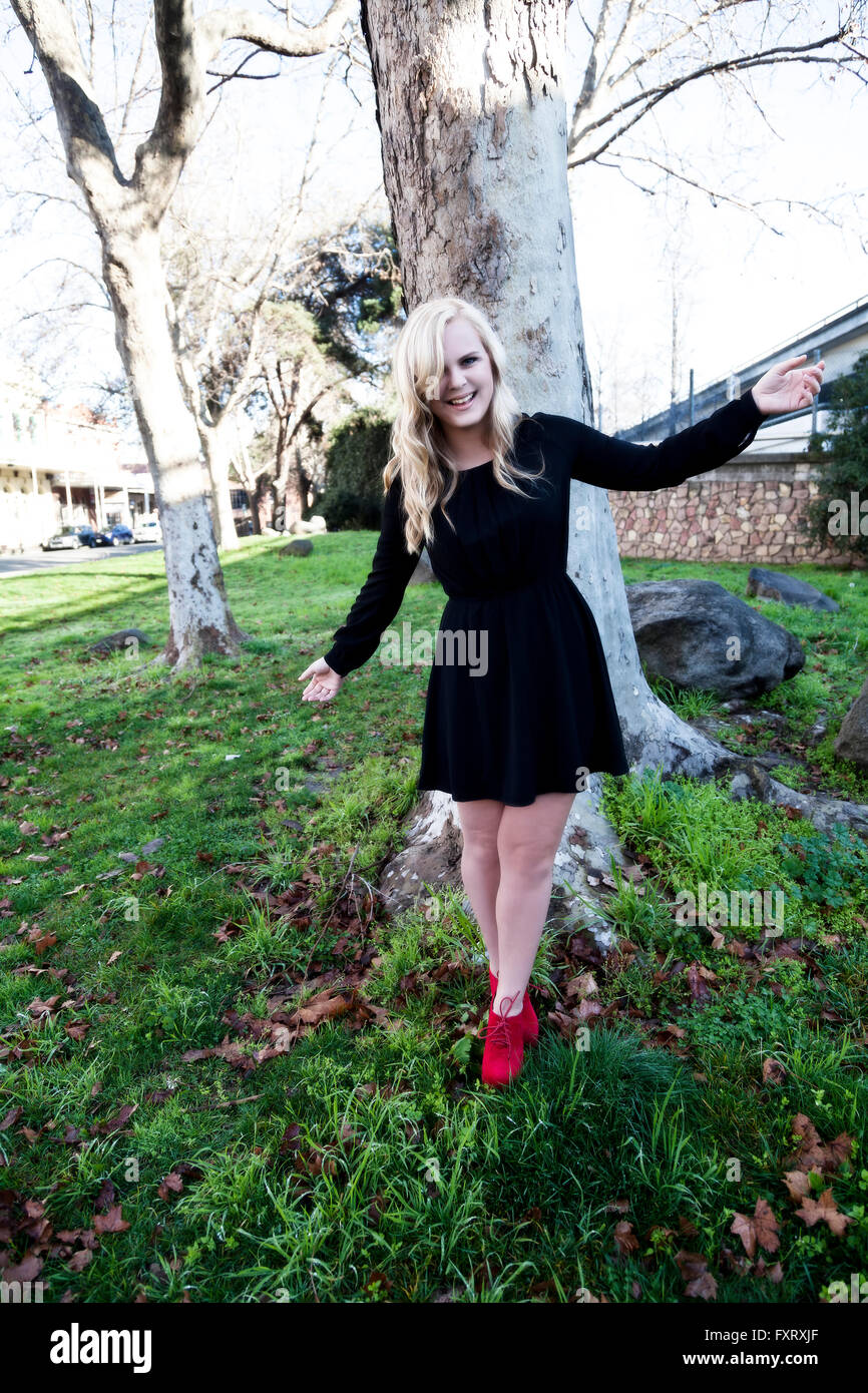 Smiling Blonde Woman Standing In robe noire et chaussures rouges en face de Tree Banque D'Images