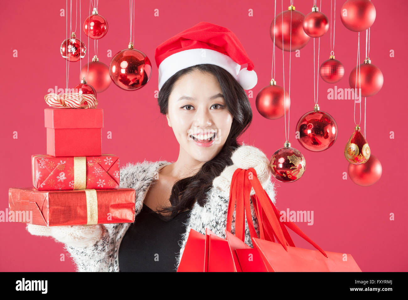 Portrait of young smiling woman wearing santa hat holding présent empilé des boîtes et sacs de magasinage à regarder devant avec des boules de Noël Banque D'Images