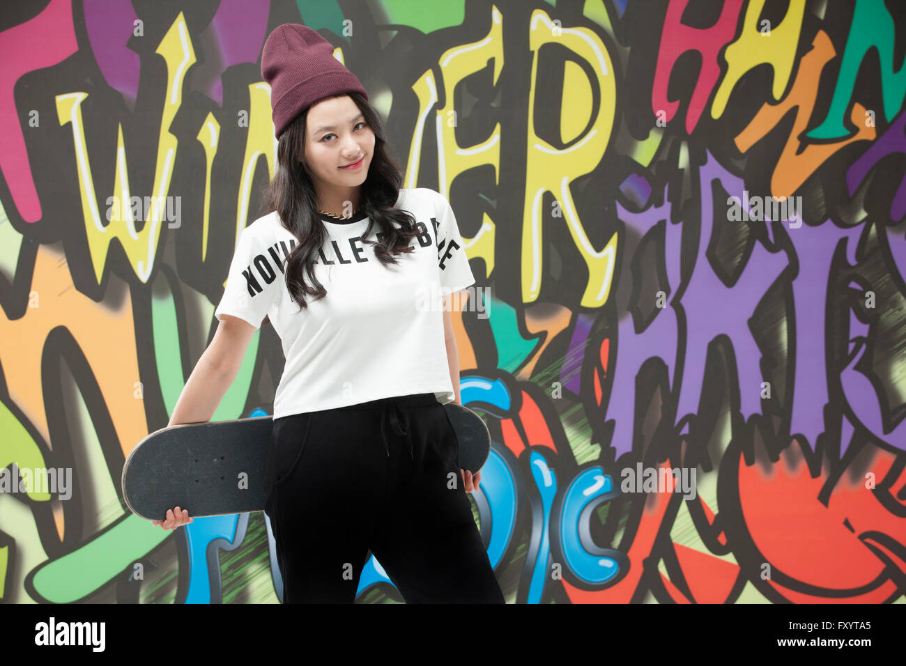 Young smiling woman in style hip-hop holding a skateboard regarder front contre l'art du graffiti Banque D'Images