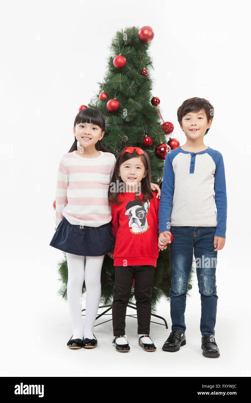 Trois enfants souriants debout main dans la main à regarder front contre l'arbre de Noël Banque D'Images
