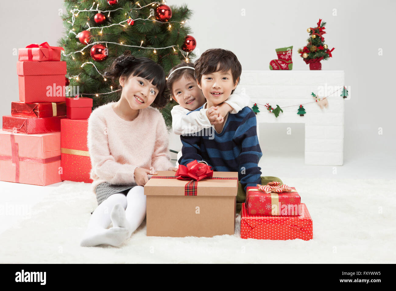 Trois enfants souriants avec boîtes actuelle fixant front contre l'arbre de Noël et cheminée Banque D'Images