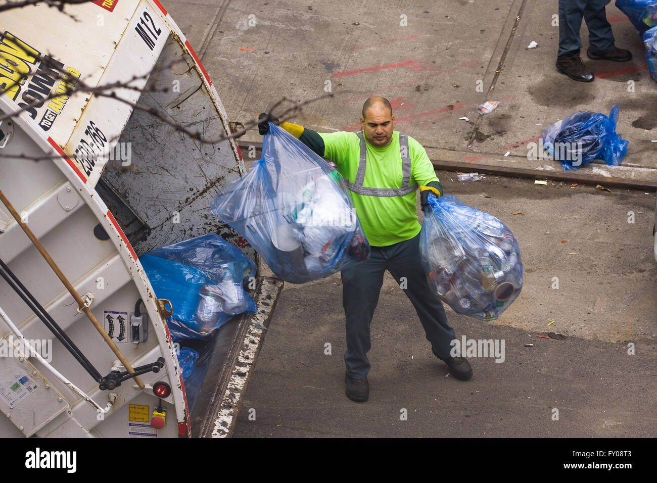 Un travailleur de l'assainissement dans la ville de New York de jeter des sacs poubelles de couleur bleue remplie de matières recyclables à l'arrière d'un camion à ordures Banque D'Images