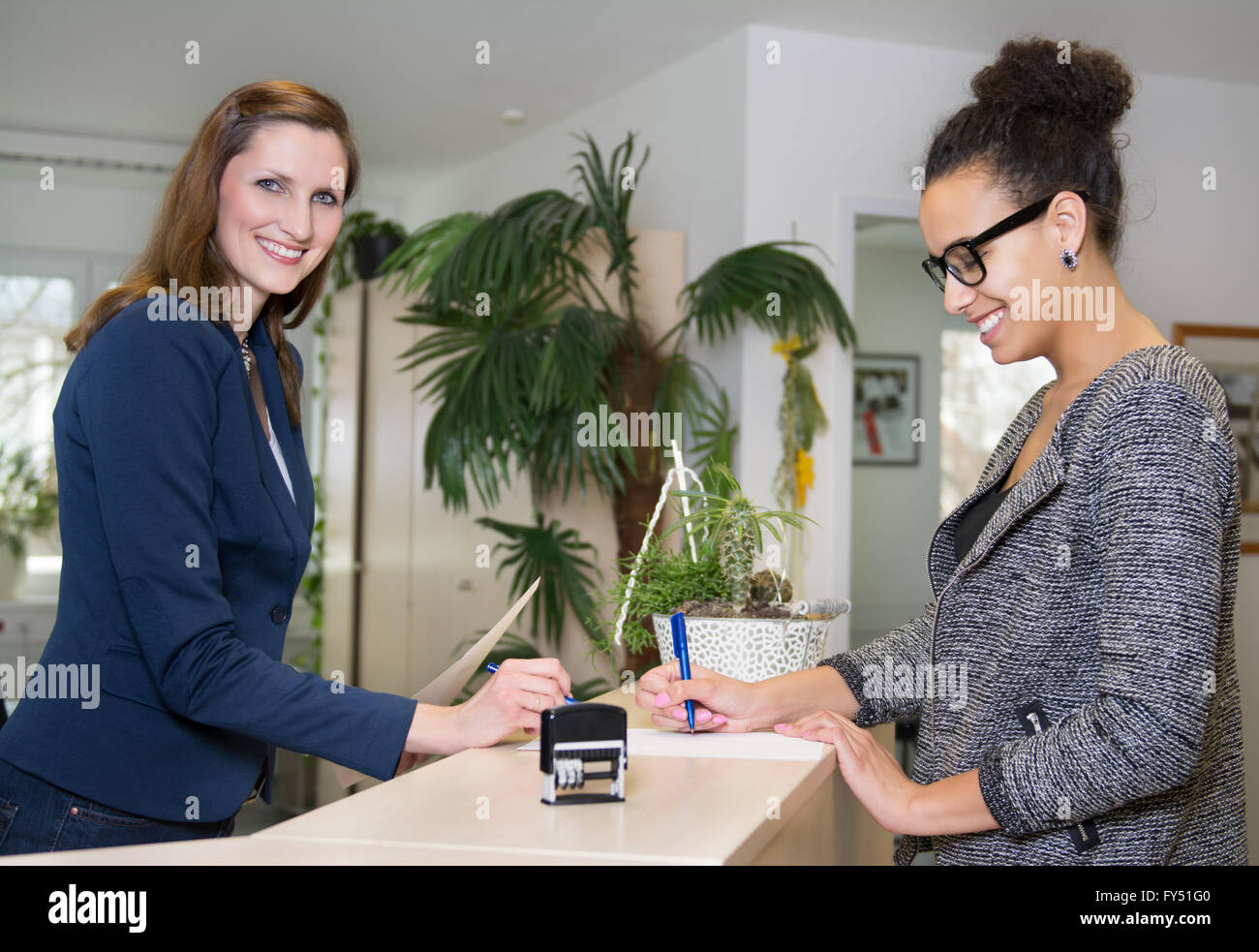 Smiling caucasian (greffier) est debout en face d'un client (latin), qui est de remplir un document à un comptoir d'accueil en th Banque D'Images