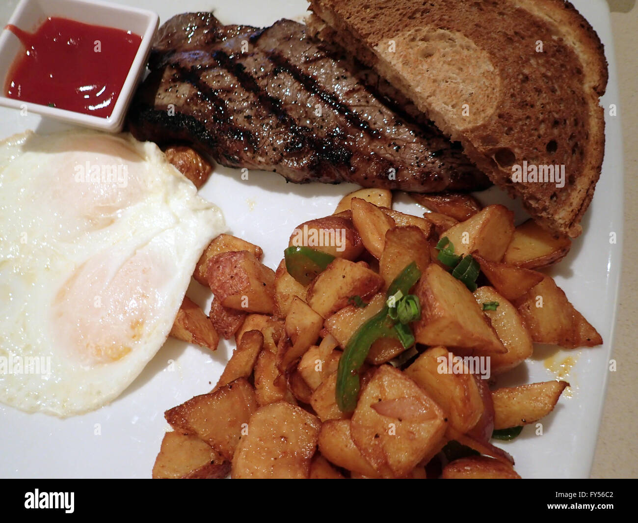 Steak avec deux œufs plus facile, deux tranches de pain de seigle, patates et tasse de ketchup sur la plaque carrée avec couteau steak sur table. Banque D'Images