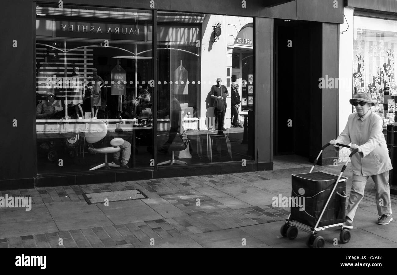 Une image en noir et blanc d'une vieille femme poussant un panier au-delà d'un magasin à la façade de verre avec de nombreuses réflexions autour de Banque D'Images