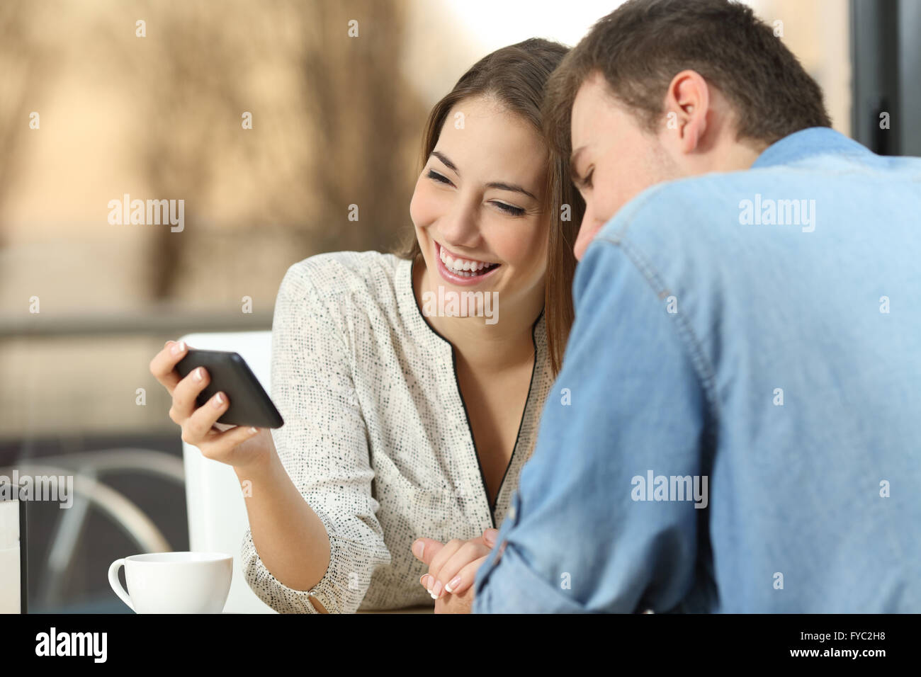 Happy casual couple rire et regarder des vidéos en streaming en ligne ensemble dans un smart phone générique dans un bar avec une ambiance chaleureuse Banque D'Images