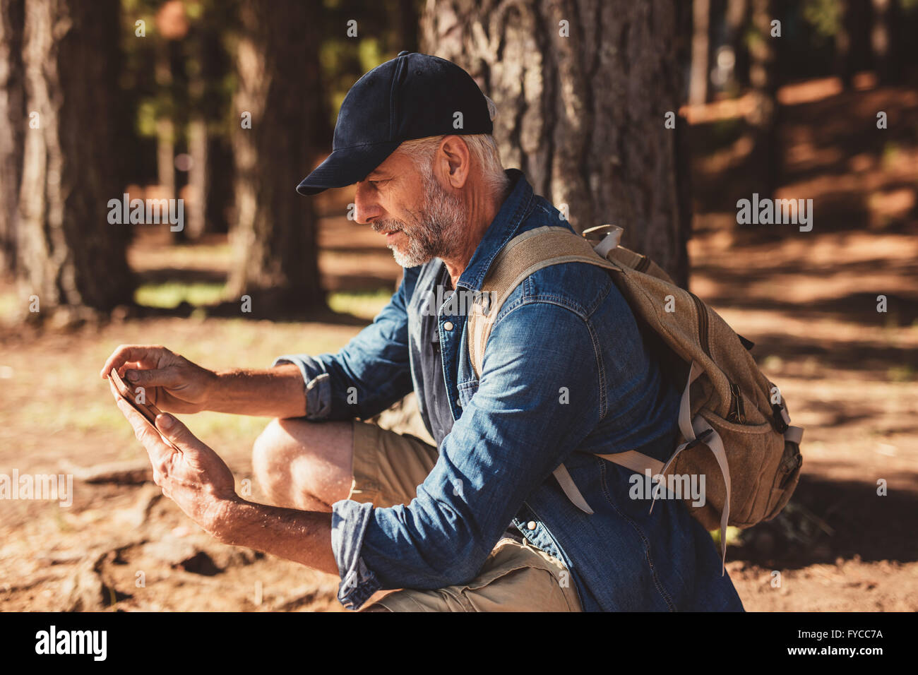 Side portrait of senior man with backpack assis dans une forêt et regardant sa tablette numérique pour les directions. Mâle mature randonnée pédestre Banque D'Images