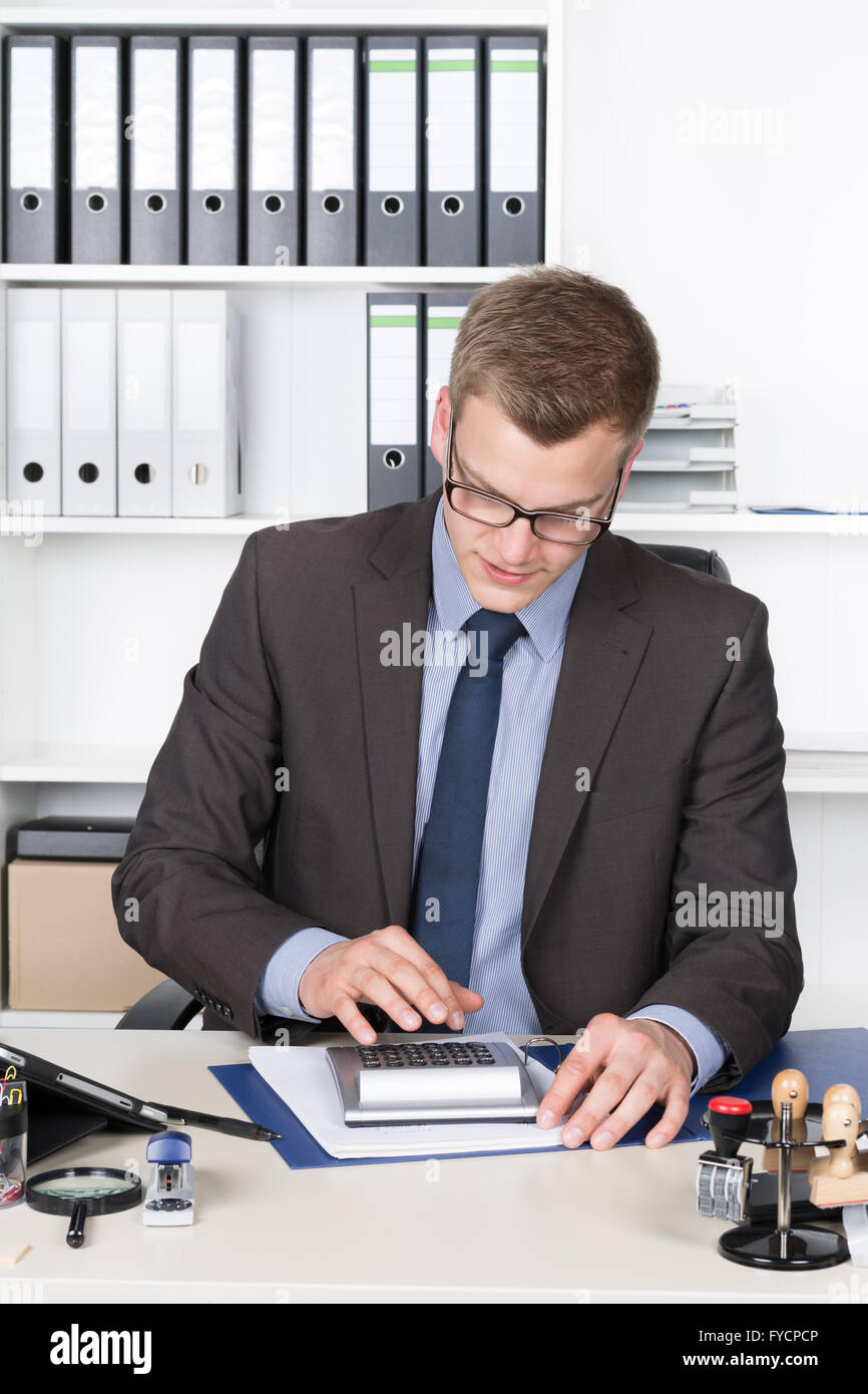 Jeune homme d'affaires est d'effectuer des calculs avec une calculatrice de bureau alors qu'il était assis au bureau dans le bureau. Une étagère est dans la b Banque D'Images
