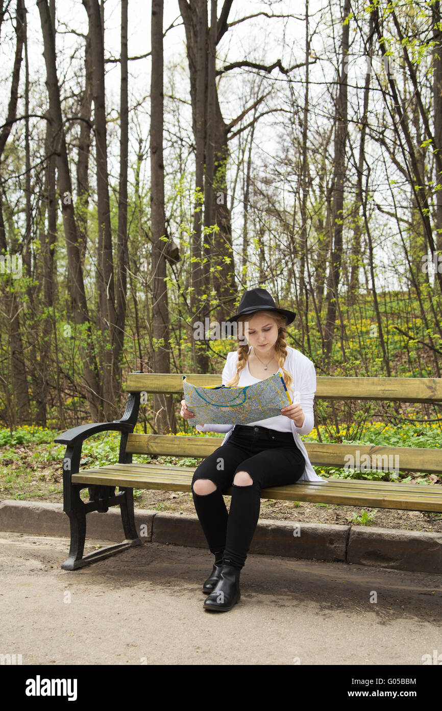 Cute blonde woman in black jeans et hat looking at map, assis sur un banc dans la forêt Banque D'Images