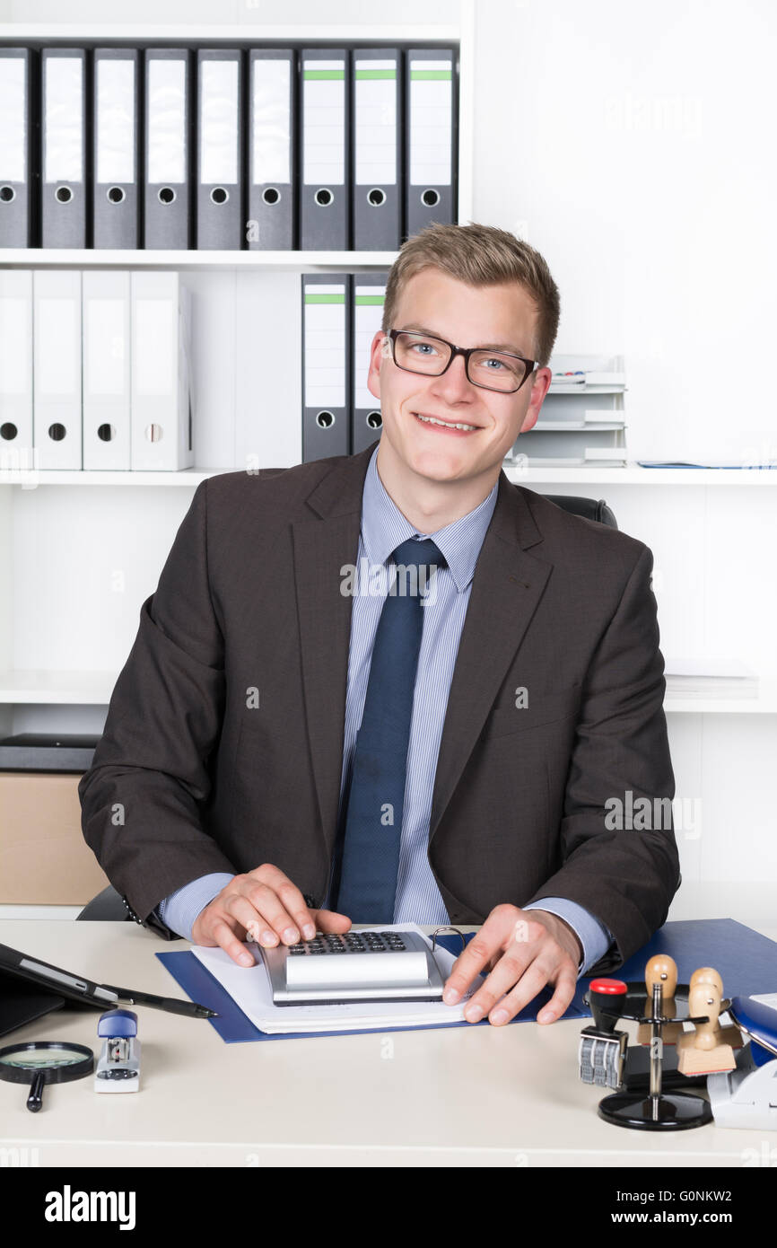 Jeune homme d'affaires est d'effectuer des calculs avec une calculatrice de bureau alors qu'il était assis au bureau dans le bureau. Une étagère est dans la b Banque D'Images