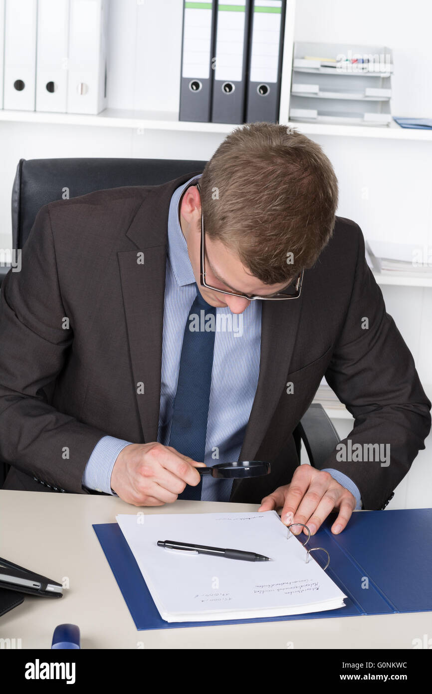 Jeune homme avec des lunettes est à la recherche d'un document grâce à une loupe alors qu'il était assis au bureau dans le bureau. Une étagère est Banque D'Images