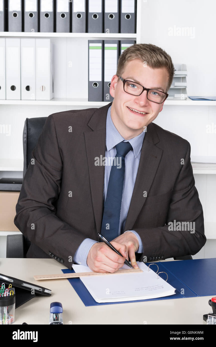 Jeune homme avec des lunettes est en utilisant une règle pour tracer une ligne dans un document alors qu'il était assis au bureau dans le bureau. Une étagère est Banque D'Images