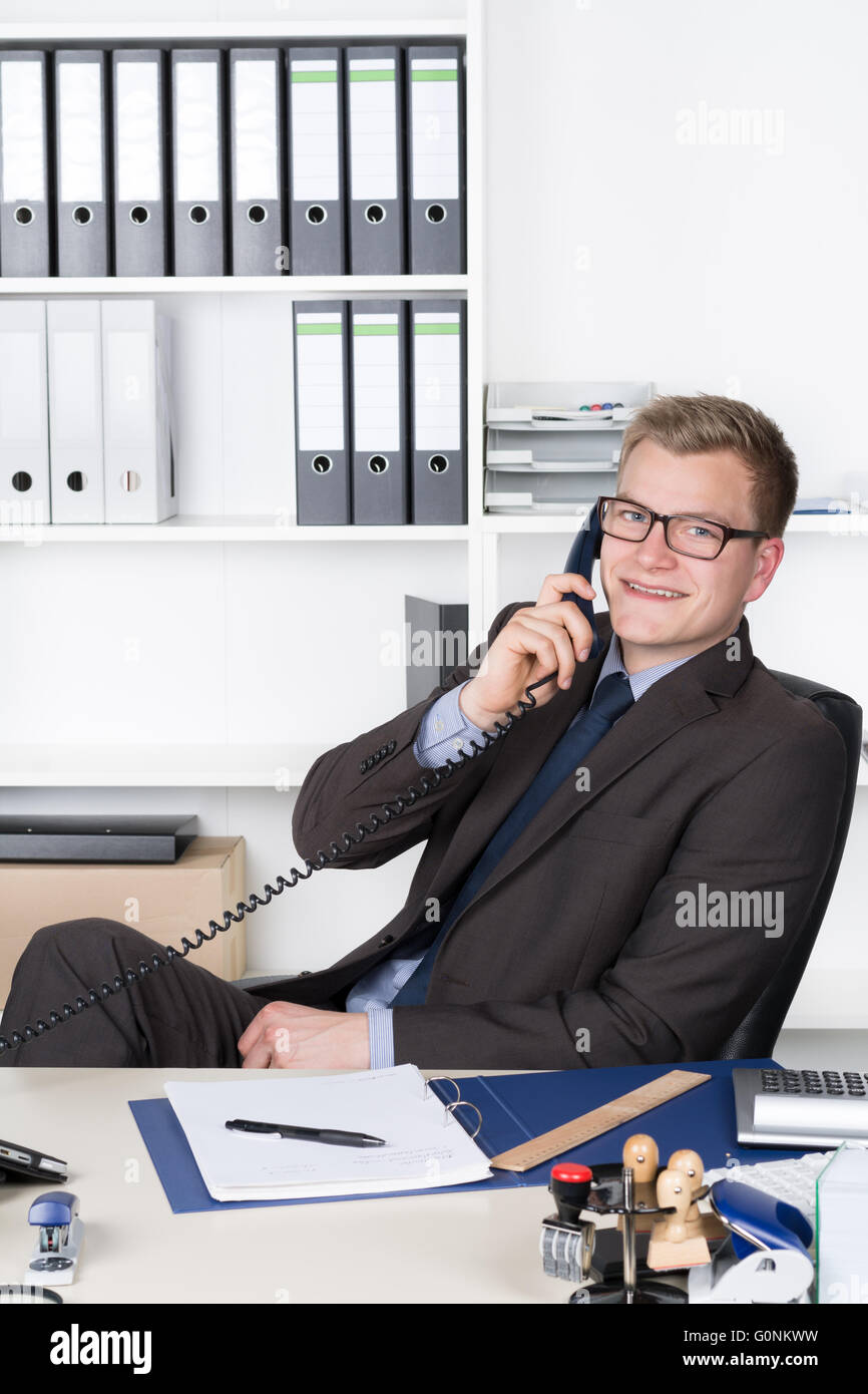 Young smiling businessman with lunettes est appelé alors qu'il était assis au bureau dans le bureau. Une étagère est dans l'arrière-plan. L'homme Banque D'Images