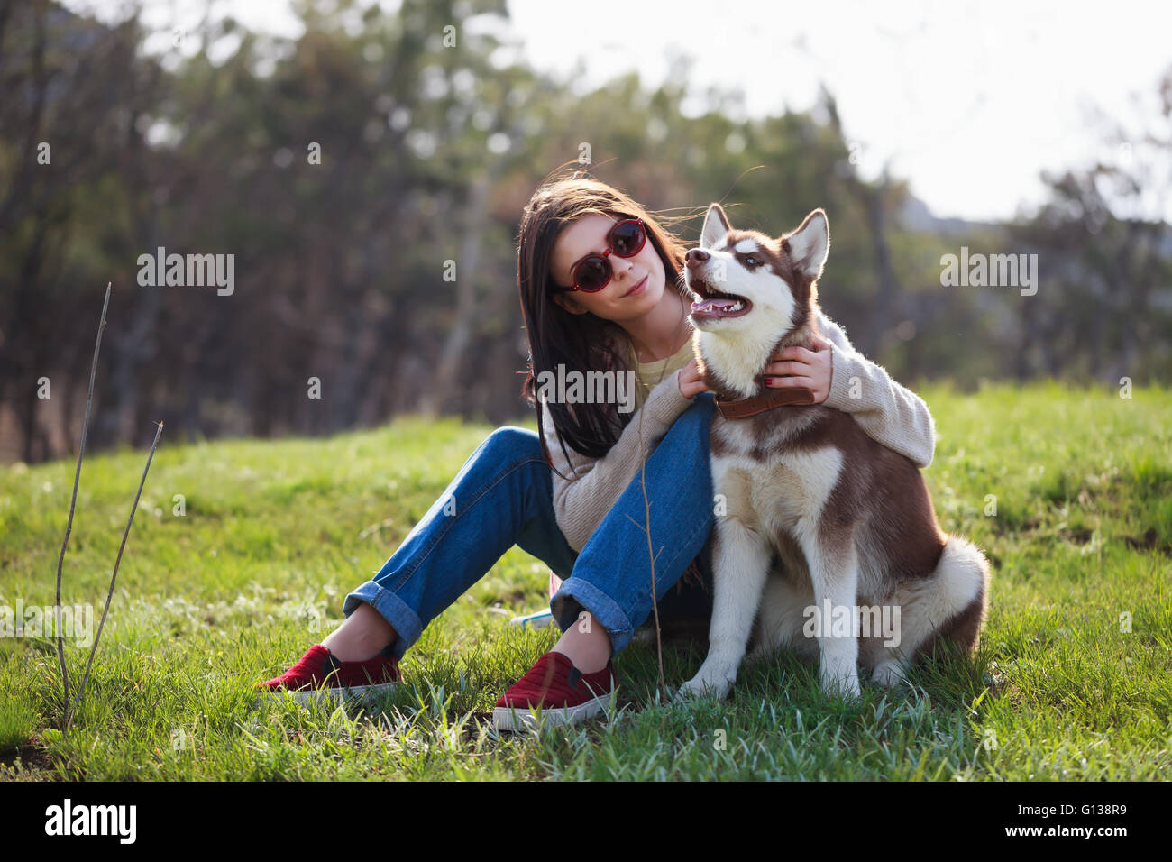Souriante jeune fille brune assise avec son chien husky dans Green Park à  l'extérieur. Cute couple sympathique et profiter de la nature. Jolie femme  et de moelleux en levrette Photo Stock -