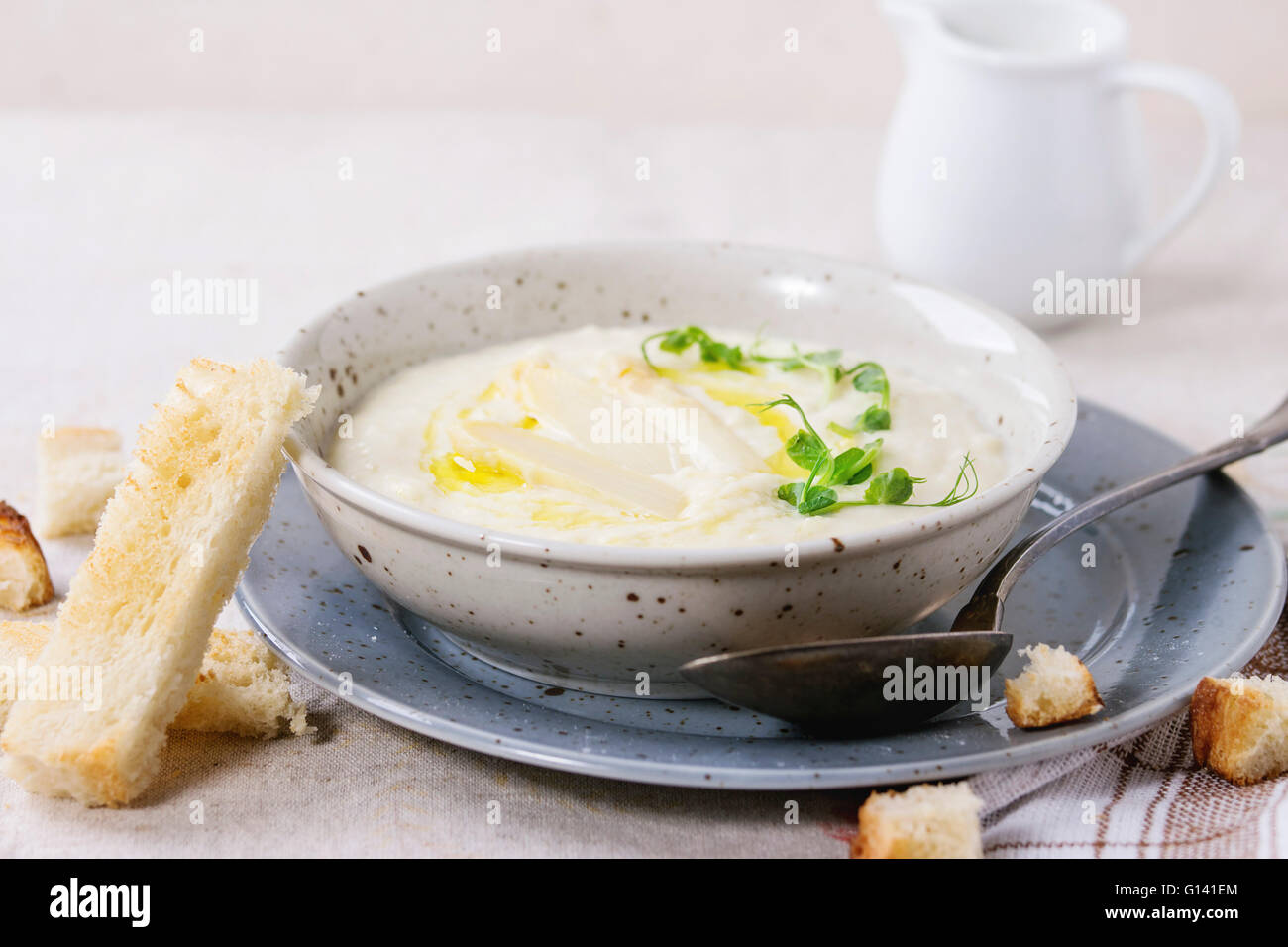 Bol de soupe crème d'asperges blanches avec des pousses de pois et de grillé, servi avec verseuse de lait sur nappe gris Banque D'Images