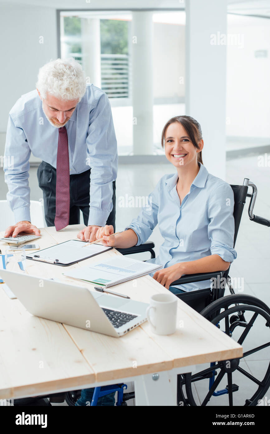 Business Woman in wheelchair travaillant au bureau avec son collègue masculin and smiling at camera Banque D'Images